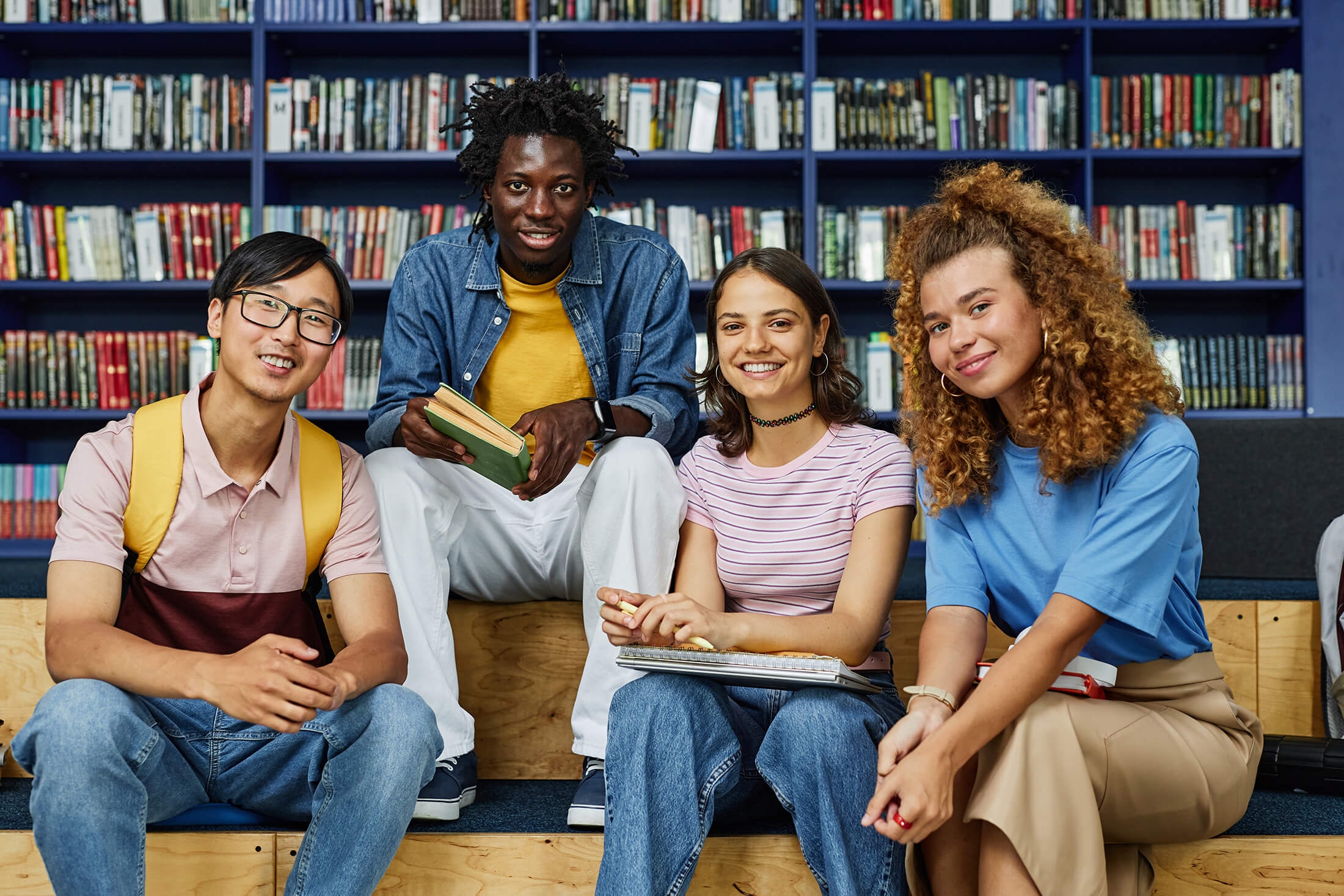 Front view at diverse group of students in library smiling happily at camera against bookshelves