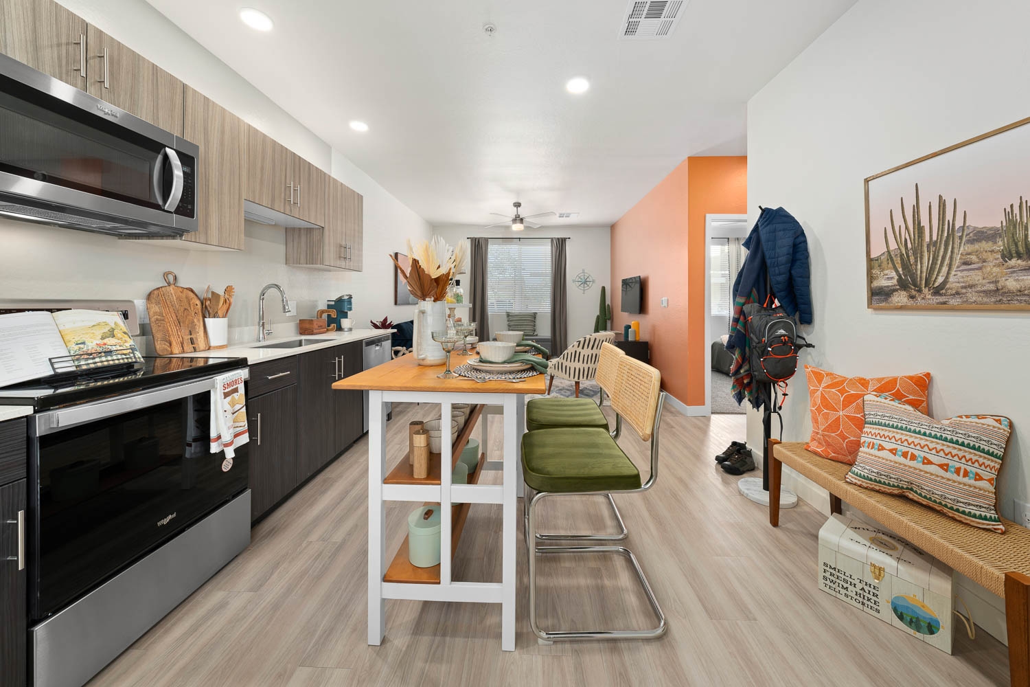 A modern apartment kitchen and living area featuring light wood flooring, a white and wood kitchen island with two green chairs,