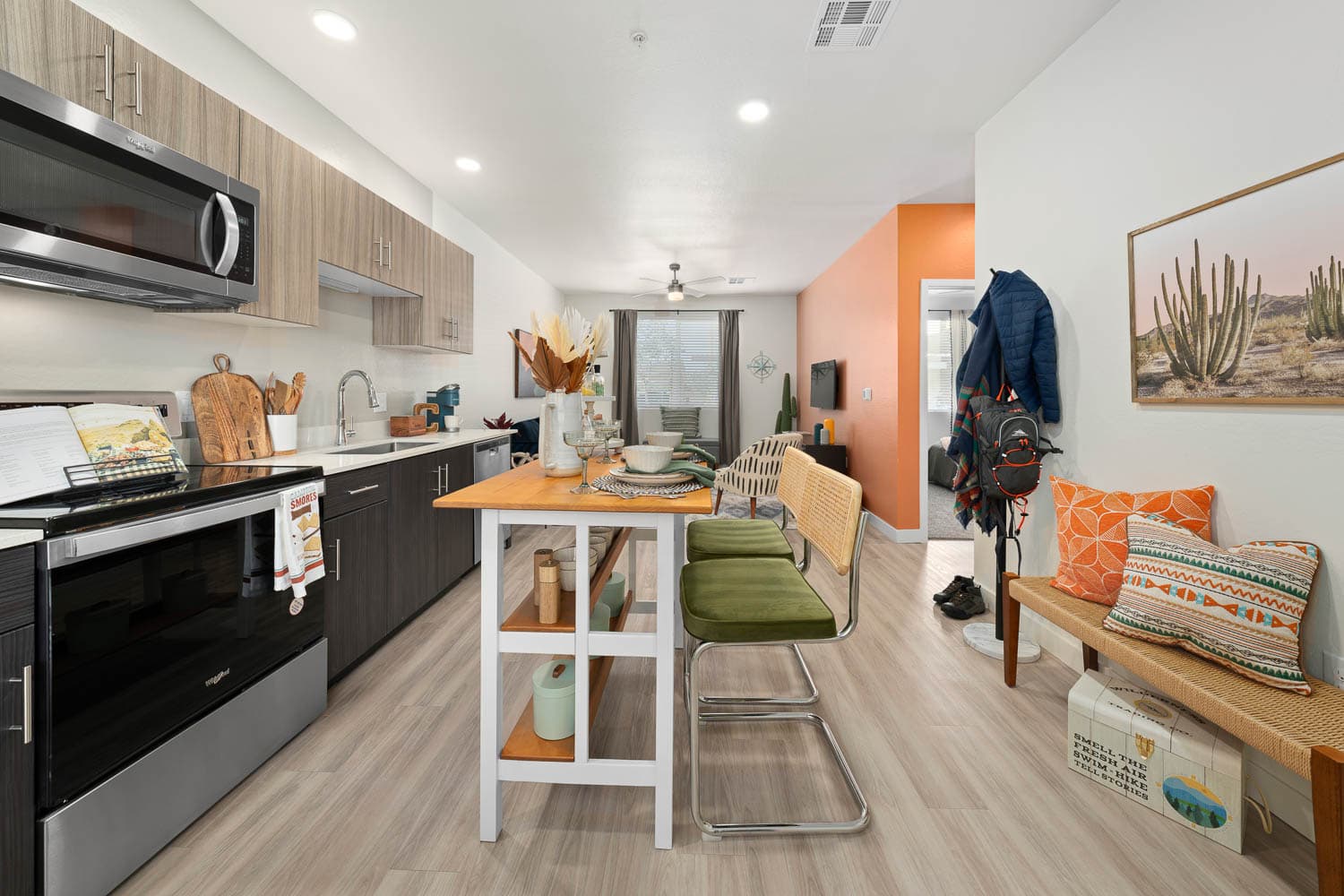 A modern apartment kitchen and living area featuring light wood flooring, a white and wood kitchen island with two green chairs,