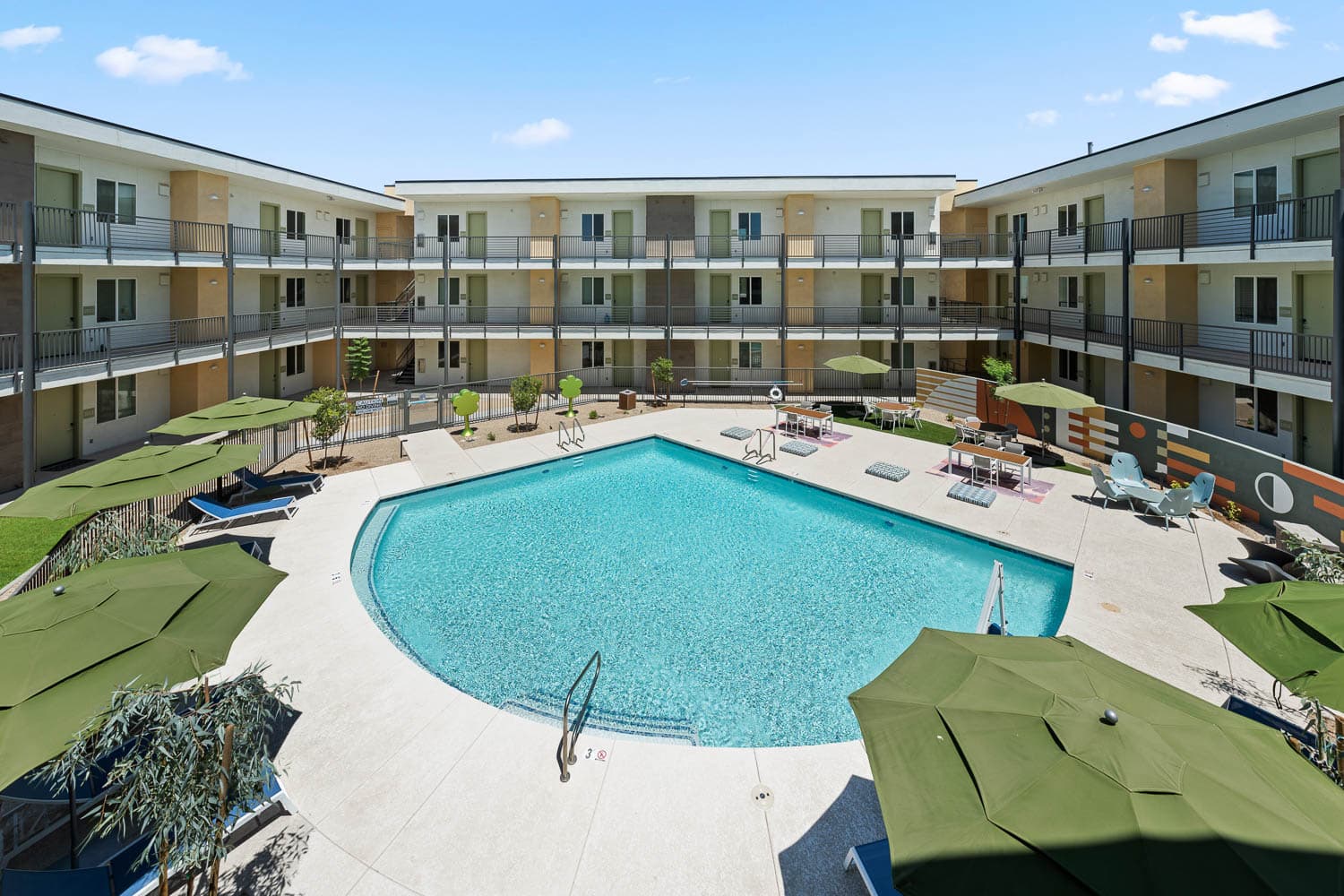 An aerial view of an apartment featuring a large swimming pool, lounge chairs with green umbrellas, and outdoor seating areas.