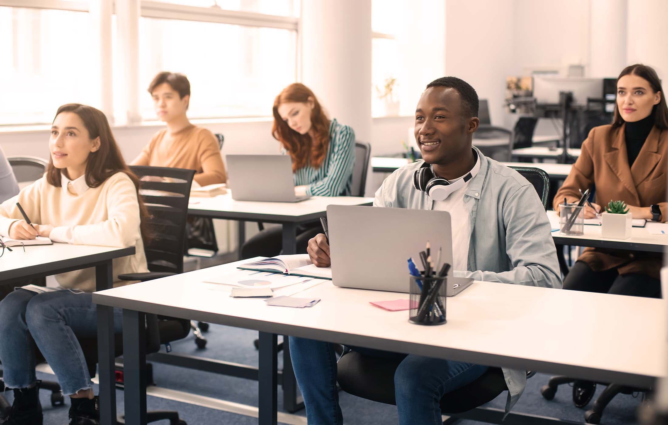 A group of students are seated, using laptops and taking notes during a class or training session.